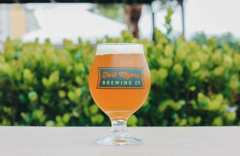 A glass of beer sitting on a table in front of bushes.